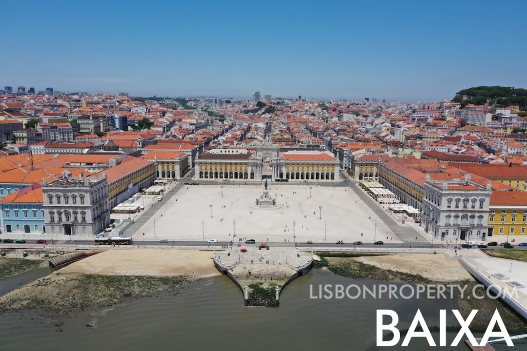 View of the Lisbon Baixa neighbourhood from the Tagus River. In the foreground, the Cais das Colunas is visible along the riverbank. Moving into the center of the scene, the Praça do Comércio and Terreiro do Paço are clearly seen, forming a significant part of Lisbon's public space. The Augusta Street Arch stands out as a notable landmark, and beyond it, the downtown rooftops of Lisbon Baixa