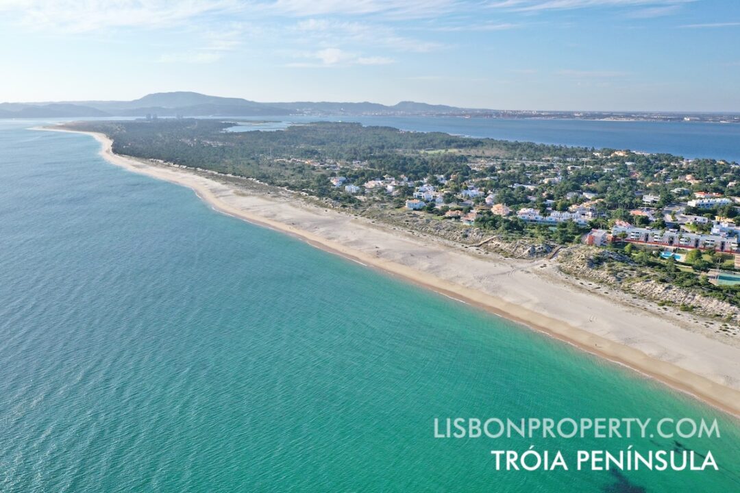 View of the Tróia Peninsula from south to north, featuring the Sol Troia tourist area with vacation villas and apartments. At the tip of the peninsula, you'll find the Tróia Resort and the marina. The Sado River is on the right, and the Atlantic Ocean is visible on the left side. On the horizon, the Arrabida Mountain Range appears on the left, while the city of Setúbal is located center-right.