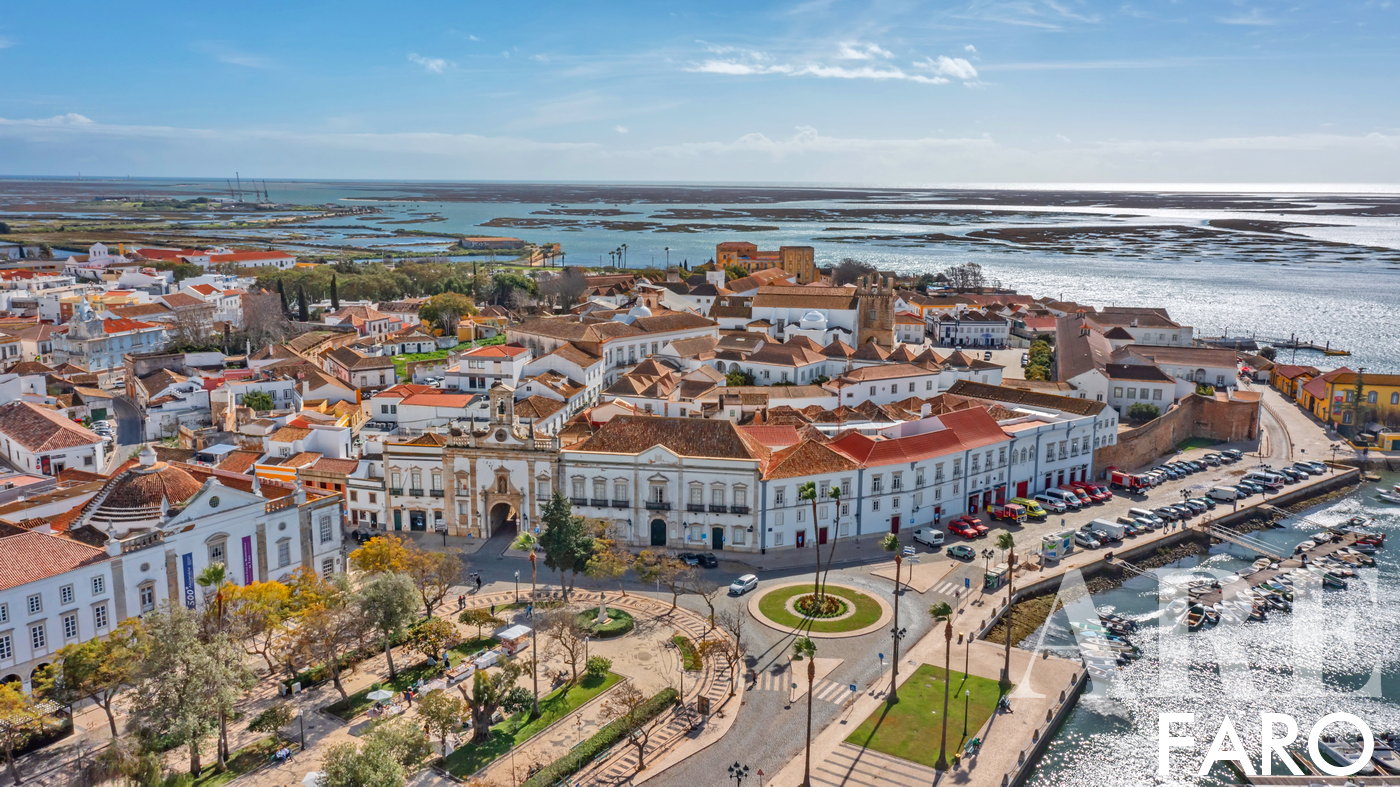 Vista aérea de Faro, coma doca no canto inferior esquerdo, o centro histórico rodeado de muralhas, a Ria Formosa e as ilhas barreira da Deserta, e o Farol no horizonte