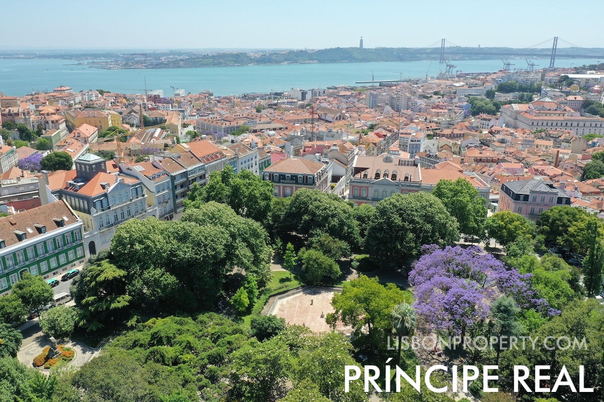 View of Principe Real garden square located at the top of the hill in the Santo António and Misericórdia parishes. The view shows the facade of buildings in the Misericórdia area with French influence from the Napoleonic period, the Tagus River, and the city of Almada on the south bank, with Cristo Rei and the 25 de Abril Bridge