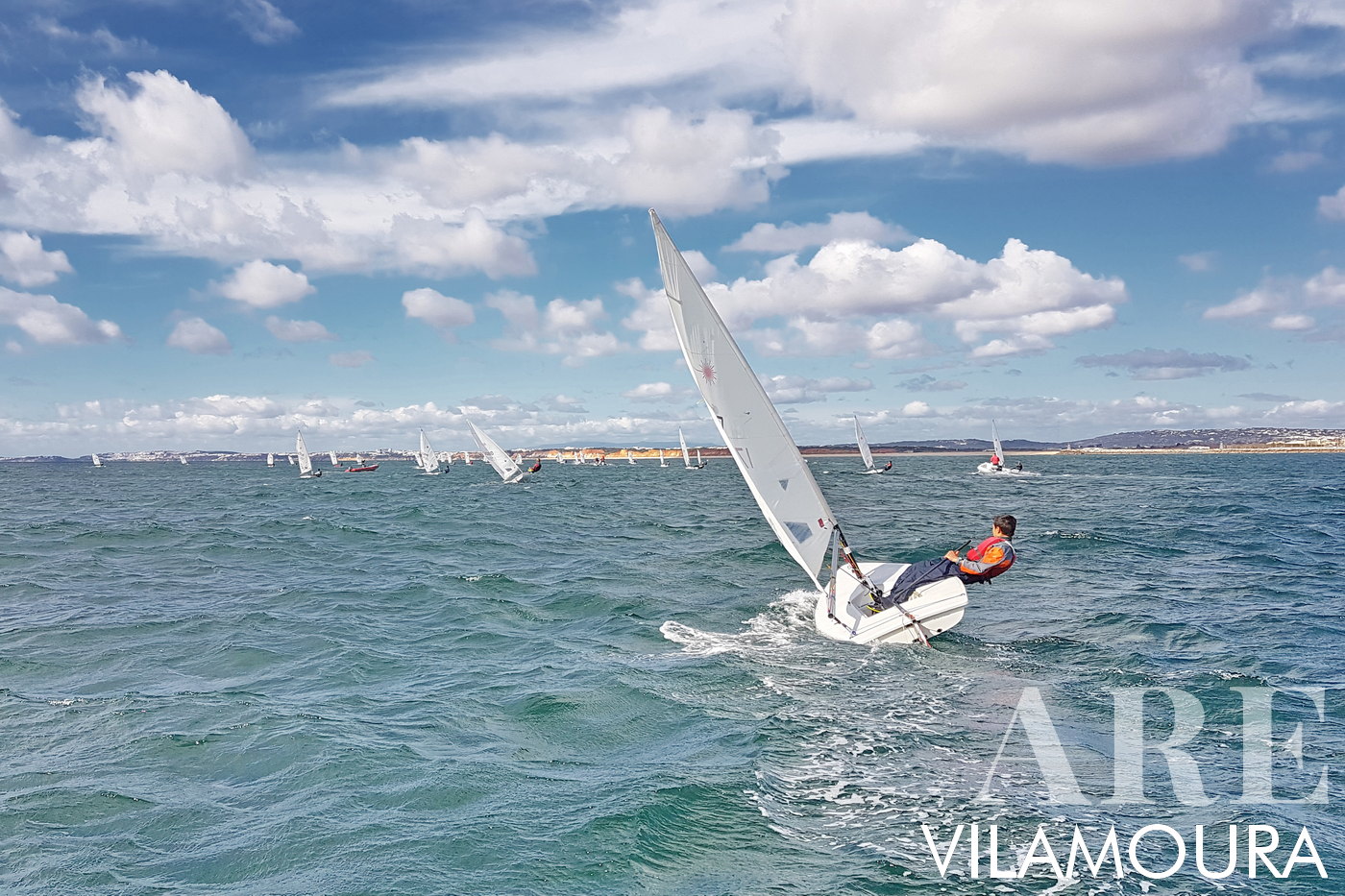 <em>Vilamoura Sailing</em><br>The captivating image vividly portrays the February Carnival Regatta at Vilamoura Marina, a time teeming with water sports enthusiasts. The focal point is a lively regatta, where young sailors expertly navigate sailboats across the glistening blue sea, their sails strikingly white against the clear sky. This spectacle is not just a race, but a testament to sailing prowess, teamwork, and a shared passion for maritime adventure. With Vilamoura