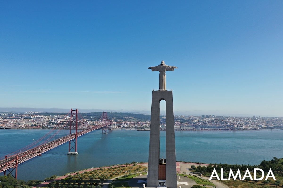This image shows a panoramic view of the Tagus River, with the city of Lisbon visible from Almada. In the foreground, the National Sanctuary of Christ the King (Cristo Rei) stands prominently, watching over the river and city.